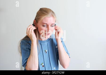 Primo piano di una donna che inserisce un auricolare wireless nell'orecchio mentre tiene il case di ricarica su uno sfondo pulito. Foto Stock