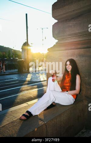 Donna elegante che si rilassa su un ponte storico della città al tramonto, godendosi l'ora dorata in un abito estivo alla moda e occhiali da sole. Foto Stock