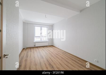 stanza vuota e luminosa con pavimentazione simile al legno. Le pareti sono bianche/grigie ruvide. Una grande finestra si affaccia su un paesaggio urbano. Una porta parzialmente visibile si trova a sinistra Foto Stock