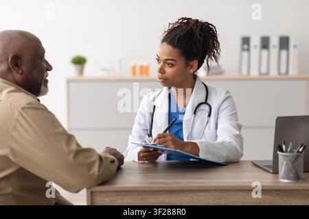 Dottore nera concentrata che consulta con un paziente maschio anziano in una clinica sanitaria Foto Stock