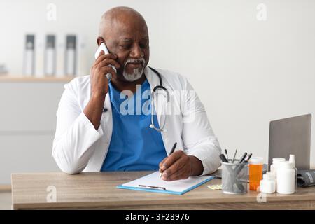 Medico uomo di colore che scrive appunti mentre parla al telefono in un ufficio sanitario Foto Stock