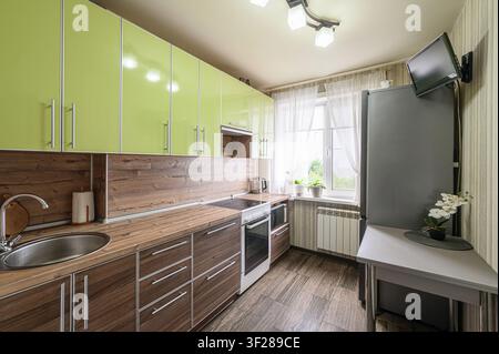 Cucina accogliente con armadi verdi, piani di lavoro in legno e un lavello in acciaio inossidabile. Una TV è montata sul muro vicino a un frigorifero Foto Stock