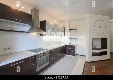 cucina moderna ben illuminata con armadi in legno scuro, piani di lavoro e piastrelle bianchi, elettrodomestici in acciaio inossidabile e pavimento in piastrelle chiare. Una grande Foto Stock