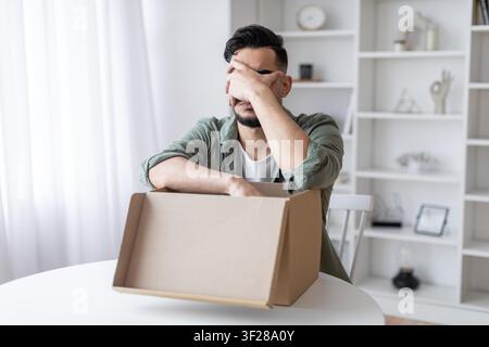 Uomo preoccupato con capelli scuri che guarda in una scatola vuota a casa, concetto di perdita di peso Foto Stock