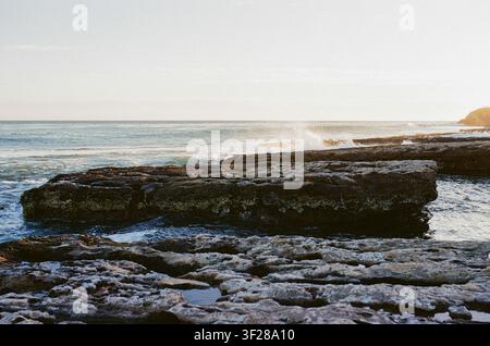 Le onde si infrangono su una costa rocciosa al tramonto Foto Stock