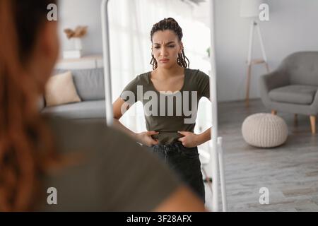 Giovane donna preoccupata che guarda il suo riflesso nello specchio, controlla l'immagine del corpo e il concetto di perdita di peso Foto Stock