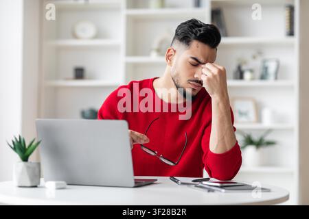 Giovane uomo preoccupato in maglione rosso che tiene occhiali mentre lavora in ufficio a casa, concetto di assistenza sanitaria Foto Stock
