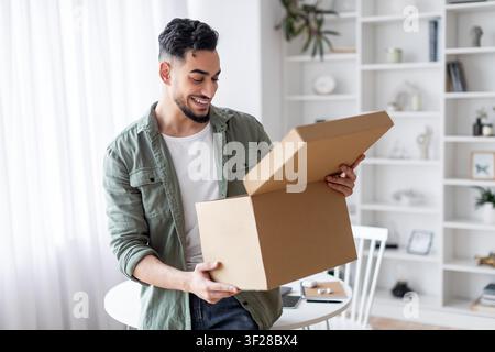 Giovane uomo felice che apre una scatola marrone a casa, sbocco dell'eccitazione, concetto di consegna Foto Stock