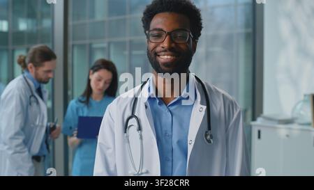 Uomo afroamericano medico che pensa, guarda la telecamera, sorride con un sorriso ampio e felice in un laboratorio clinico vicino a lavoratori della medicina sullo sfondo. Uomo etnico Foto Stock