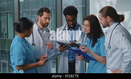 Cinque medici multietnici concentrati con tablet digitale che lavorano, parlano, decidono la diagnosi medica nel corridoio della clinica. Infermiere e praticanti multirazziali Foto Stock