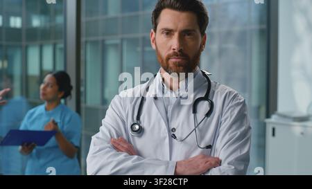 Ritratto di un uomo caucasico serio e sicuro di sé, medico, che posa con le mani incrociate in ospedale. Terapeuta maschile autorevole in uniforme medica bianca che guarda Foto Stock