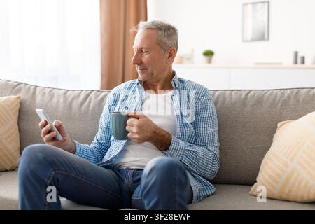 Uomo caucasico anziano che si rilassa sul divano con telefono e caffè a casa, concetto di stile di vita felice Foto Stock