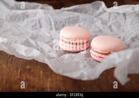 Macarons rosa teneri su carta, sfondo di legno. Bacche di bosco, sapore di fragola o lampone Foto Stock