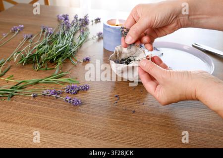 Mani umane che riempiono una busta di lino con fiori di lavanda essiccati, sfondo tavolo di legno. Candela accesa, mazzo di fiori viola freschi, vista laterale Foto Stock