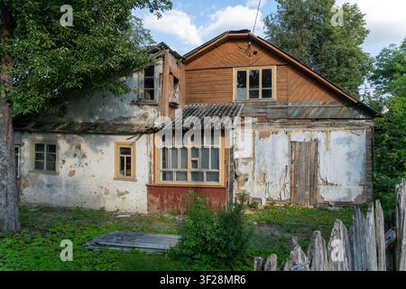 Vecchia casa di legno fatiscente e abbandonata con vernice scrostata e finestre rotte. Giardino invaso dalla vegetazione e recinto consumato in un ambiente rurale. Foto Stock
