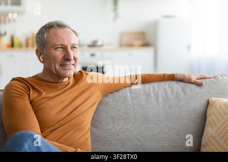 Uomo caucasico anziano sorridente che si rilassa a casa sul divano, ascoltando musica con gli auricolari, soggiorno Foto Stock