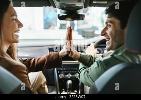 Giovane uomo e donna felici che si divertono con un gesto di high five in macchina, sorridendo insieme Foto Stock