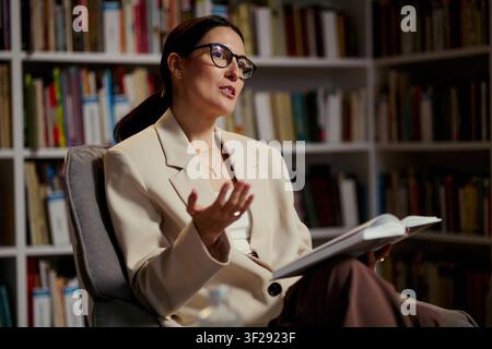 Donna caucasica di mezza età che parla e gesticola mentre tiene un libro aperto in ufficio, trasmettendo consulenza o coaching professionale. Utile per terapia, consulenza, marketing educativo Foto Stock