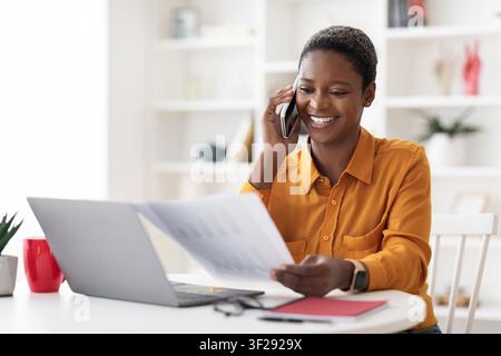 Donna afroamericana sorridente al telefono mentre lavora alla scrivania in ufficio, concetto aziendale Foto Stock