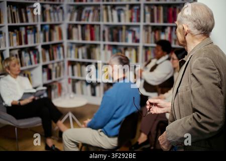 Uomo anziano in piedi e partecipa a una discussione di libro con un gruppo adulto diversificato in biblioteca, condividendo la sua prospettiva e imparando attivamente. Utile per l'educazione, la comunità, le campagne di alfabetizzazione Foto Stock