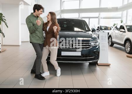 Giovane coppia felice che celebra l'acquisto di una nuova auto in sala espositiva, emozione gioiosa Foto Stock