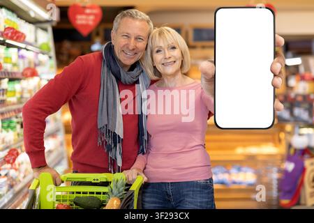 Coppia anziana caucasica felice che fa la spesa insieme al supermercato, tiene uno smartphone con schermo vuoto Foto Stock
