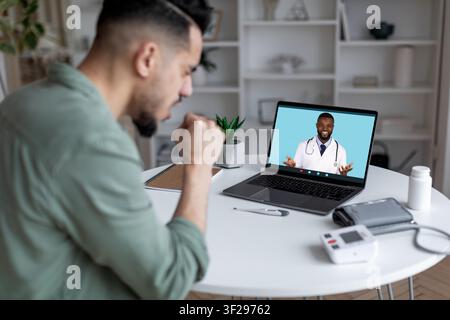 Uomo che pensa e consulta con un medico nero sullo schermo a casa, concetto di assistenza sanitaria Foto Stock
