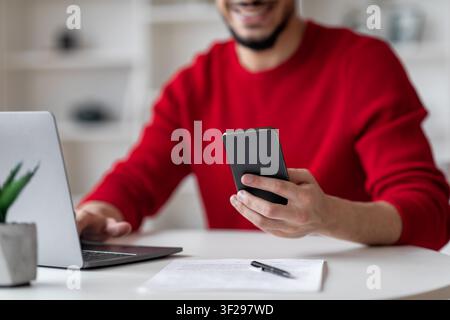 Giovane uomo sorridente che tiene uno smartphone mentre lavora a una scrivania in un ufficio moderno, concetto aziendale Foto Stock