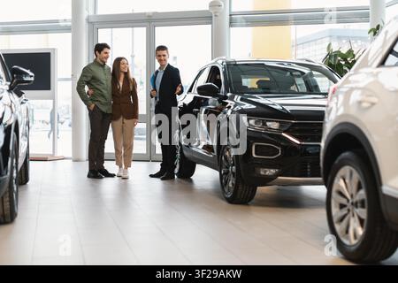 Coppia che discute delle opzioni dell'auto con il venditore nella sala espositiva, felice e coinvolta, concetto aziendale Foto Stock