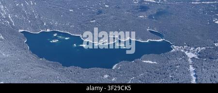 Un colpo aereo panoramico ad angolo alto del profondo lago blu dell'Eibsee con le sue piccole isole, completamente circondato da una fitta foresta di pini innevata in Foto Stock