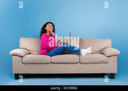 Giovane donna afroamericana che lavora su un laptop, pensa mentre si rilassa sul divano, concetto di casa Foto Stock