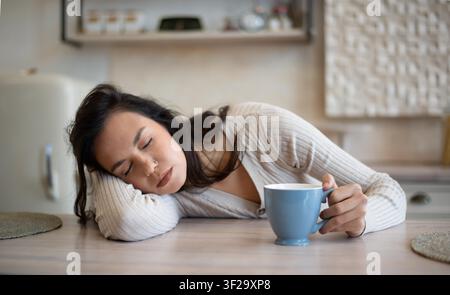 Giovane donna caucasica stanca che dorme sul tavolo con una tazza di caffè in cucina, concetto di relax Foto Stock