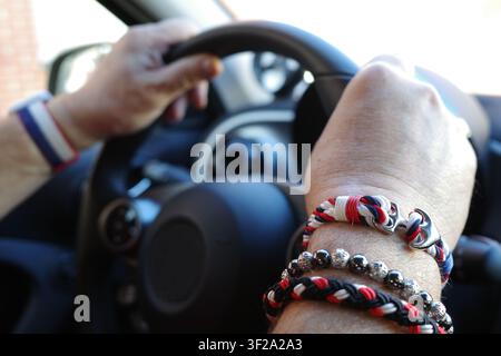 Mani che tengono un volante di auto, adornate con più braccialetti in rosso, bianco e blu. I braccialetti sono una combinazione di intrecciati e perlati des Foto Stock