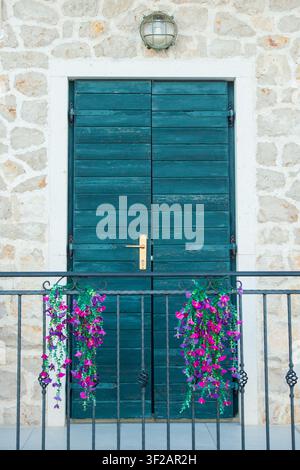 Una porta di legno blu rustica inserita in un muro di pietra con una luce esterna vintage e una ringhiera del balcone decorata con fiori vivaci viola e rosa. Foto Stock