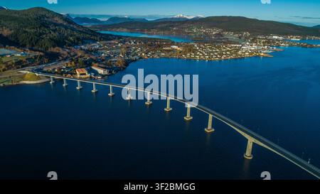 Vista aerea di un lungo ponte che si estende su un profondo fiordo blu, circondato da colline ondulate, case costiere e cielo sereno, Norvegia. Foto Stock