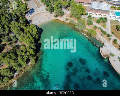 Vista aerea che mostra acqua turchese chiara e una spiaggia di ciottoli circondata da rigogliosi pini verdi e infrastrutture di resort costiero Foto Stock