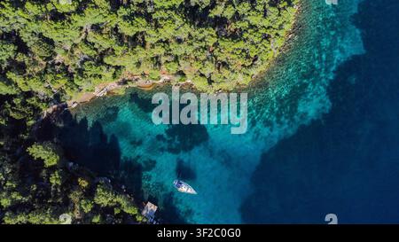 Vista aerea che mostra una costa boschiva verde con uno yacht a vela ancorato nelle acque turchesi chiare e blu profonde di una baia isolata Foto Stock