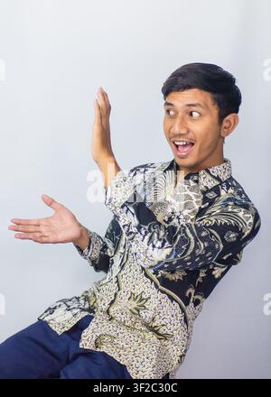 Giovane asiatico sorpreso con camicia in batik che guarda la fotocamera con espressione scioccata, isolato su sfondo da studio Foto Stock