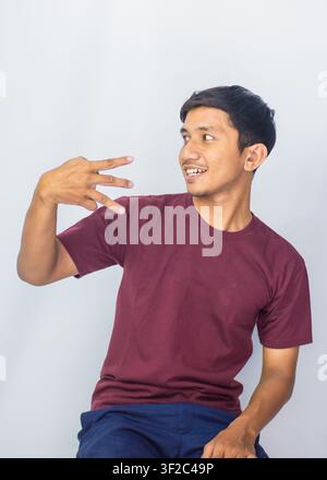 Giovane asiatico allegro in polo viola che fa il gesto della vittoria su uno sfondo blu in studio. Foto Stock