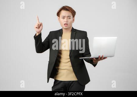 Ritratto di un uomo d'affari asiatico arrabbiato che tiene un computer portatile, indossa un completo e fa un gesto con il dito puntato su sfondo isolato Foto Stock