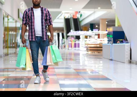 Uomo africano irriconoscibile che cammina nel centro commerciale con un sacco di borse di carta Foto Stock