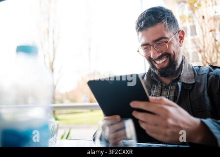 Uomo che sorride e ride guardando contenuti su un tablet digitale in un caffè Foto Stock