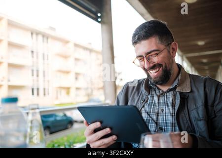 Uomo che ride guardando contenuti su un tablet digitale in un caffè all'aperto Foto Stock