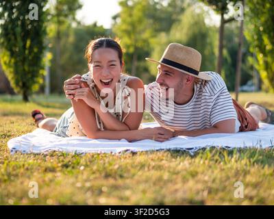 Una coppia allegra si gode un tranquillo picnic in un parco panoramico, sorridendo mentre parlano e si rilassano su una coperta. Foto Stock