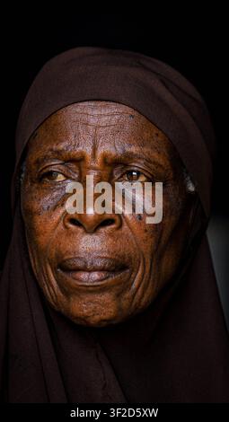 Ritratto di una donna musulmana anziana con un hijab marrone, il suo viso illuminato dolcemente su uno sfondo scuro. Foto Stock