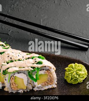 Rotoli di sushi freschi ricoperti di salsa cremosa, ripieni di avocado e verdure, serviti con wasabi vivace. Piatto nero e bacchette migliorano l'elegante presentazione. Foto Stock