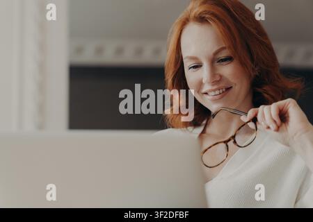 Una donna dai capelli rossi sorride mentre lavora con un laptop e tiene gli occhiali a casa Foto Stock