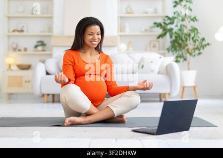 Donna nera incinta sorridente segue una sessione di meditazione online su laptop Foto Stock
