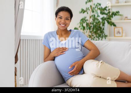 Donna nera incinta felice che si rilassa sul divano e tiene il ventre Foto Stock