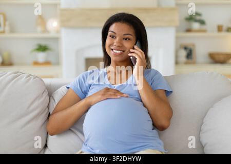 Donna nera incinta sorridente che parla al telefono mentre è seduta sul divano Foto Stock